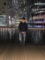 Diego standing by a wooden-deck railing at Canary Wharf at night, with glass office towers and the dock behind him.